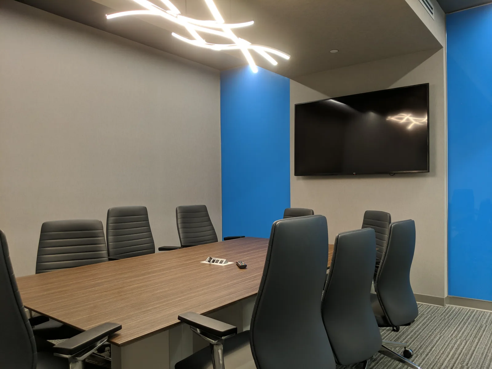 Conference room with wood table, black chairs, wall-mounted display, blue accents.