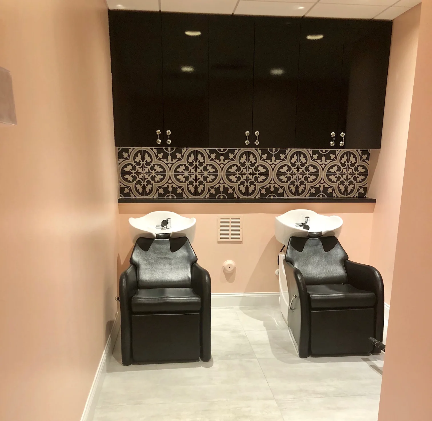 Hair salon shampoo station with two black chairs and decorative tile backsplash.